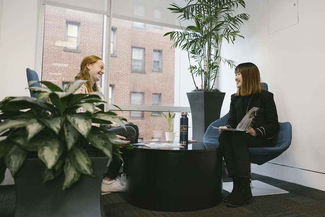 Two GWSB international business students chat at a table surrounded by plants in a student lounge in Duques Hall