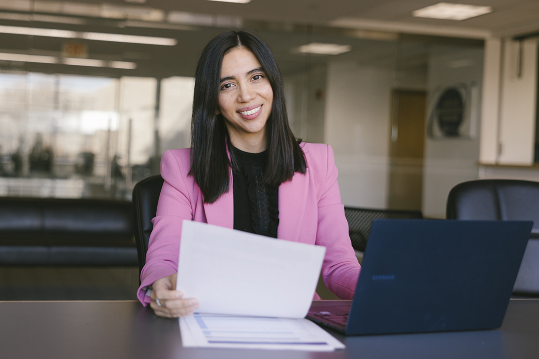 A GWSB accountancy student wearing a pink blazer smiles and sits at a table in a student lounge in Duques Hall. In her hand, she holds a piece of paper.